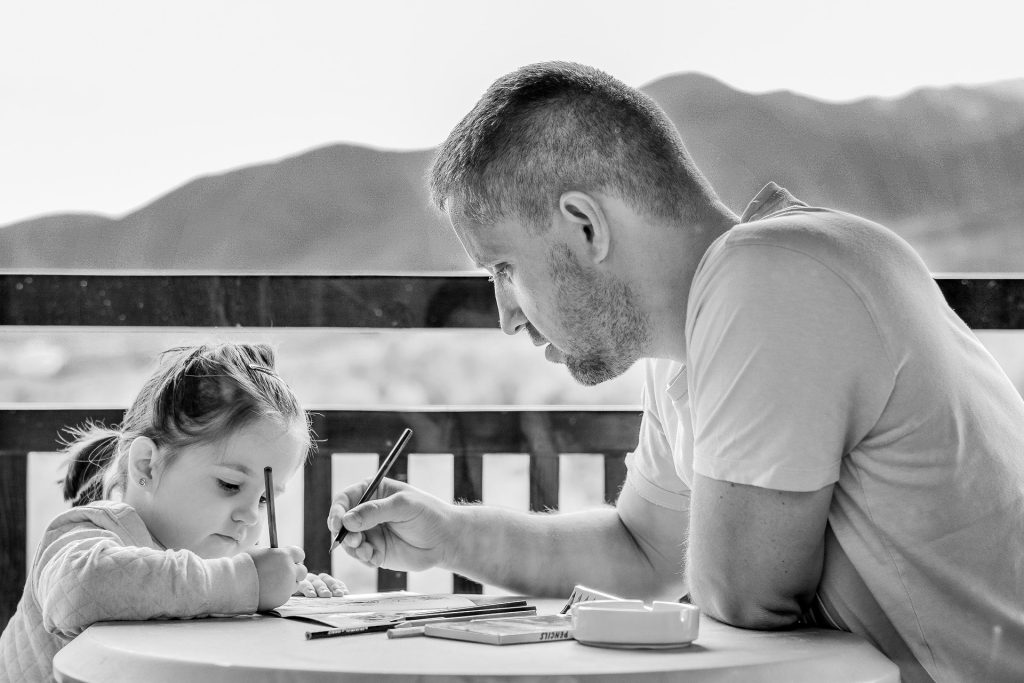 An adult and a young child sit at a round table, drawing with pencils during a visitation. Mountains and a railing can be seen in the background.