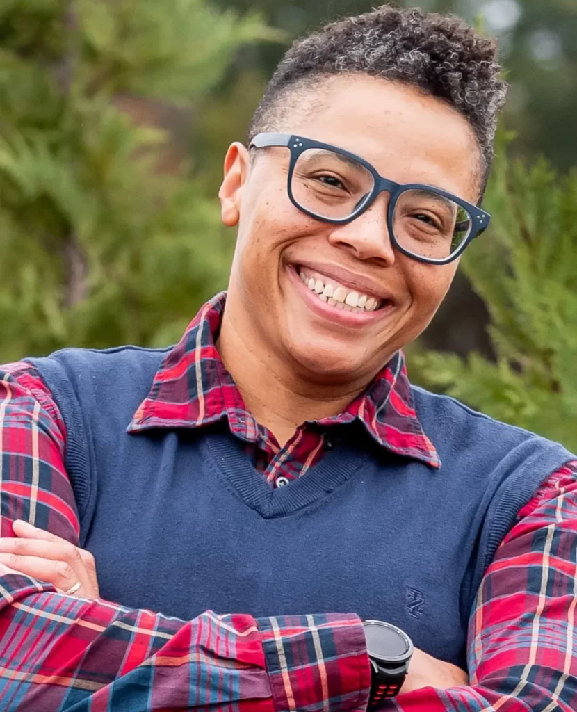 Person wearing glasses, a red plaid shirt, and a blue sweater vest smiles outdoors with green foliage in the background, radiating the warmth and approachability you’d expect from someone experienced in family law.