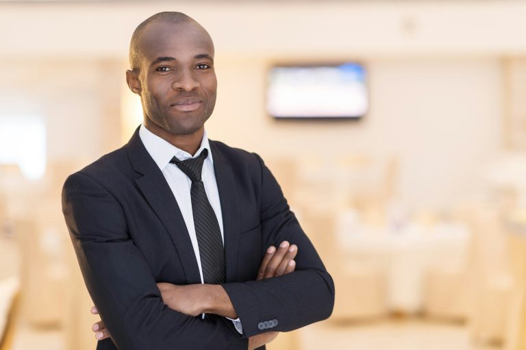 A man in a black suit and tie stands indoors with his arms crossed, looking at the camera. The background is blurred, showing a bright room with a television—a professional image often seen among African American family law attorneys.