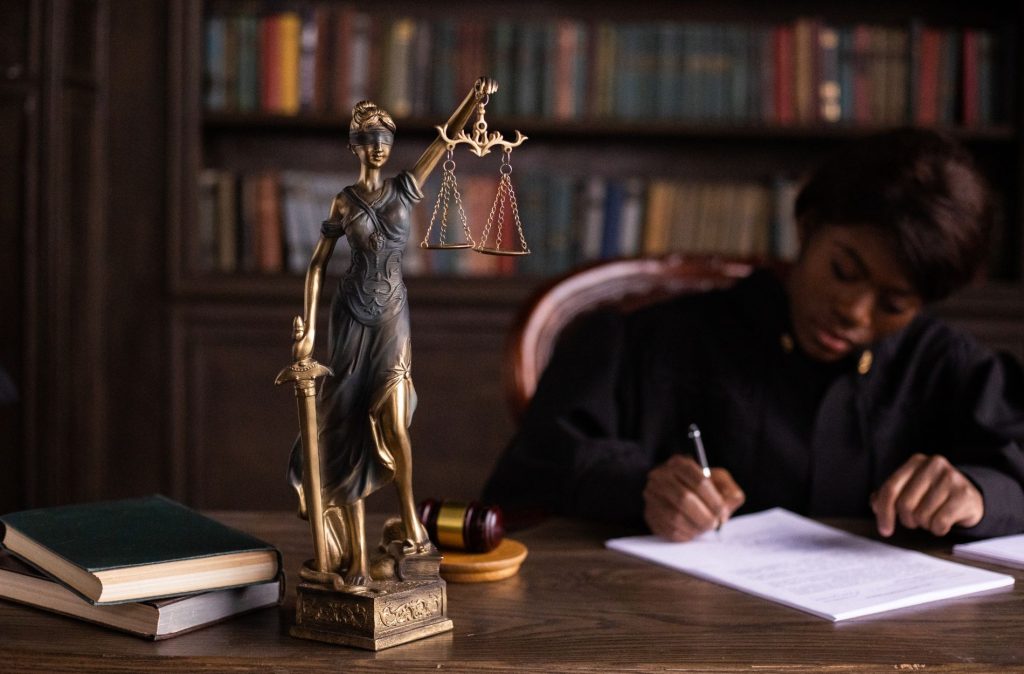 A bronze Lady Justice statue stands on a desk with books and a gavel; a person in a robe writes on papers, possibly about parent visitation or visitation schedules, with shelves of books behind them.