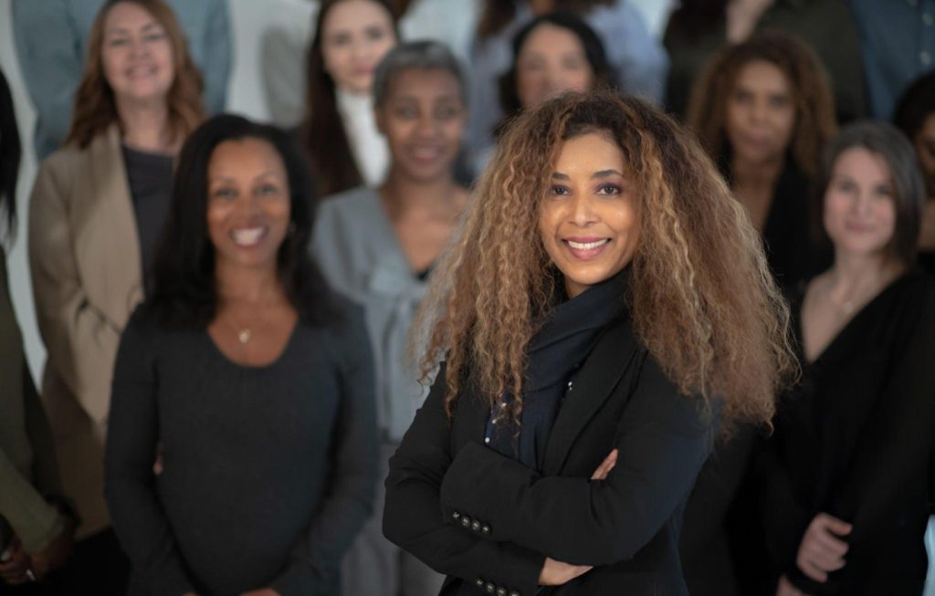 A Black female divorce attorney with curly hair stands confidently with her arms crossed in front of a diverse group, highlighting the impact on cases led by strong representation.