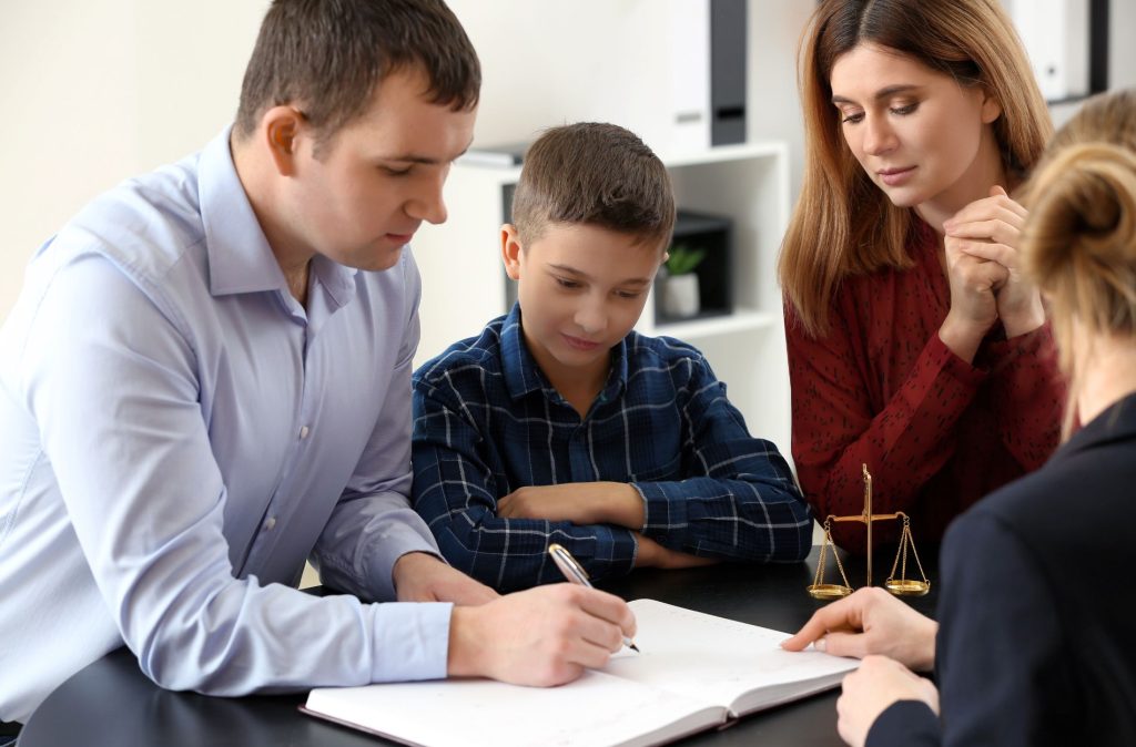 A Board Certified Family Lawyer assists as a man signs a document at a desk, accompanied by a woman, a child, and another woman; a scale of justice rests on the table.