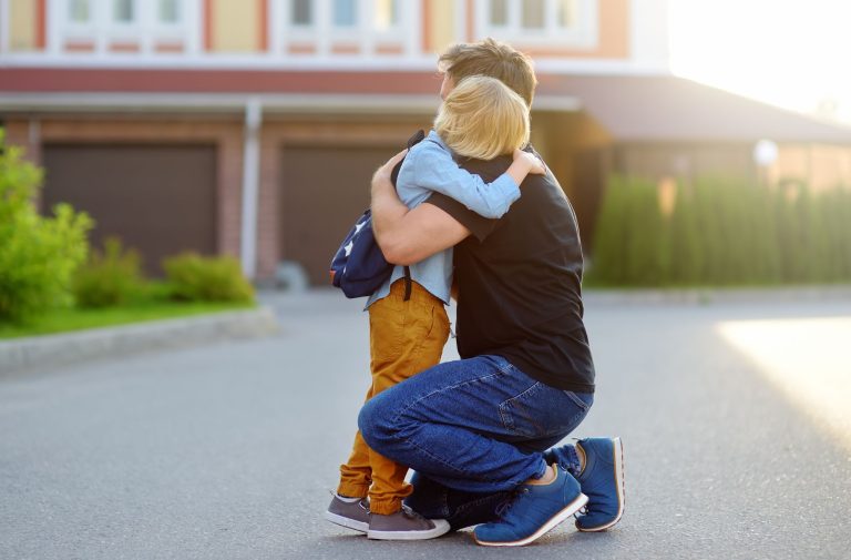 An adult and a child with a backpack hug each other outdoors in front of a house with garage doors, symbolizing the emotional journey involved in filing child custody and taking important child custody steps.