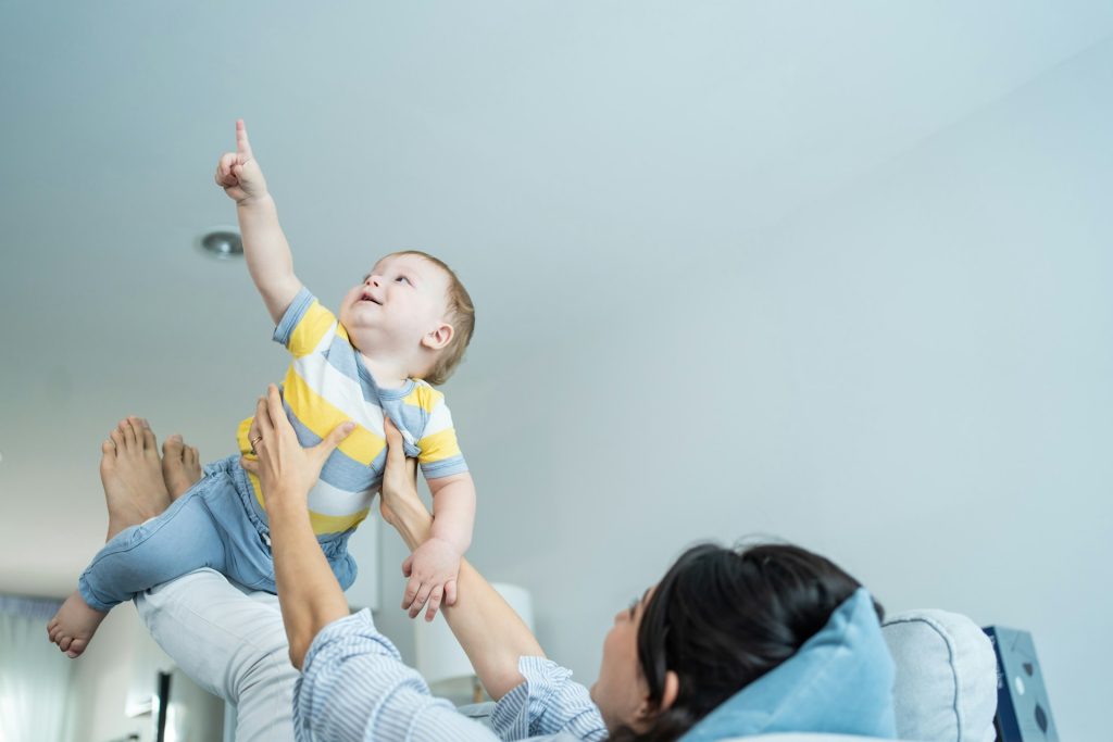 An adult lying on a couch lifts a smiling baby in the air; the baby, wearing a yellow and gray striped shirt with blue pants, points upward—a joyful moment that reminds us of the importance of child custody and cherishing every moment together.