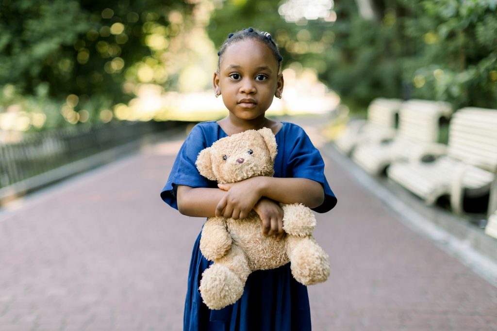 A young girl in a blue dress stands on a park path, holding a brown teddy bear with both arms—capturing the innocence at the heart of many child custody cases handled by board-certified family lawyers. Benches and trees fill the background.