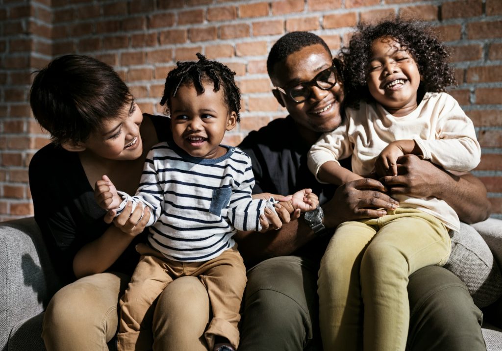 A family of four sits on a couch in front of a brick wall, smiling and laughing together—reflecting the joy that can be found with guidance from compassionate family law attorneys.