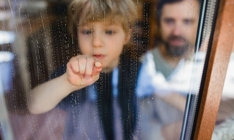 A child involved in child custody touches a rainy window from inside, with an adult standing in the background and droplets visible on the glass.