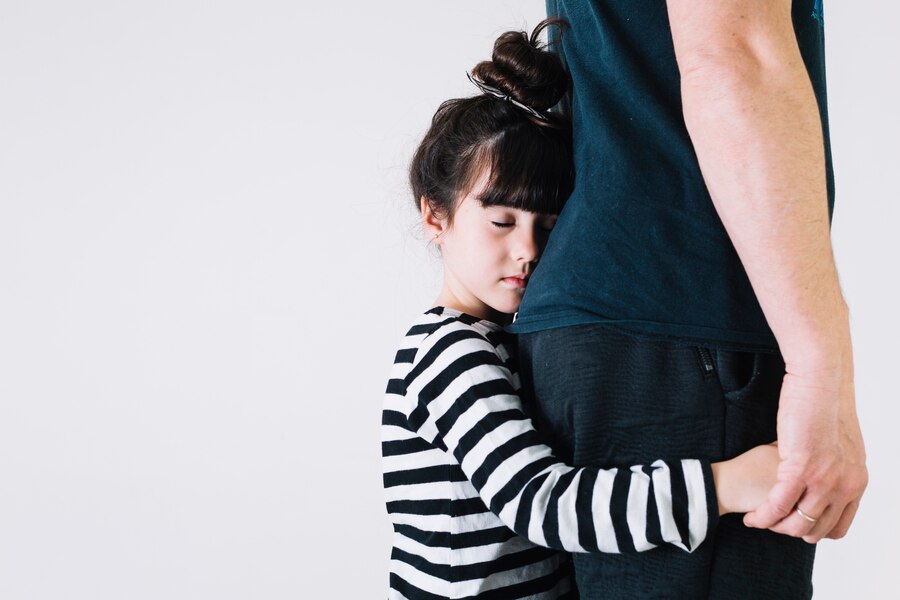 A young girl with closed eyes hugs an adult's waist from the side—a tender moment that reflects the emotional bonds at the heart of Texas child custody decisions. Both stand against a plain white background, holding hands.