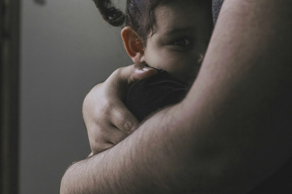 A child with dark hair and pigtails is being held closely by an adult, looking out from behind the adult's arm—a touching scene often seen during transitions in Texas custody agreements.