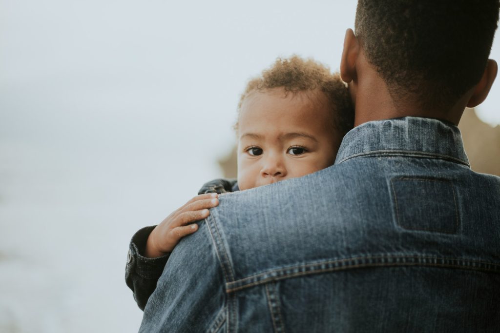 A young child rests their head on an adult's shoulder, both wearing denim jackets, with the adult facing away from the camera—capturing a tender moment that reflects the importance of fathers' rights.