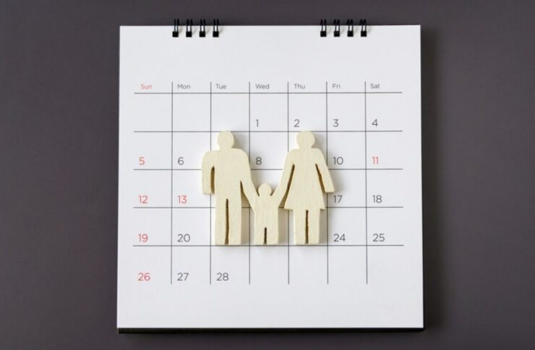 A wooden cutout of a family of three is placed on a white calendar with dates marked in red and black, symbolizing a custody calendar for effective co-parenting.