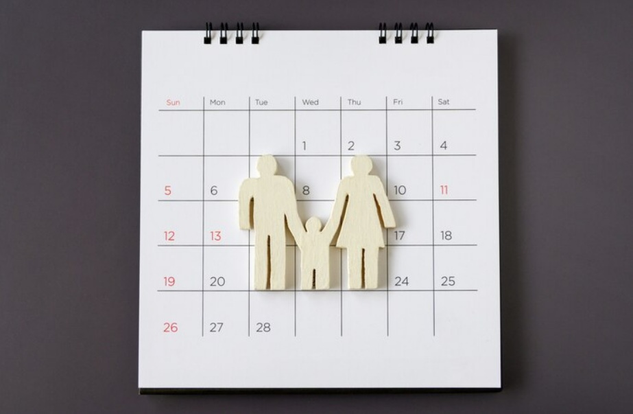 A wooden cutout of a family of three is placed on a white calendar with dates marked in red and black, symbolizing a custody calendar for effective co-parenting.