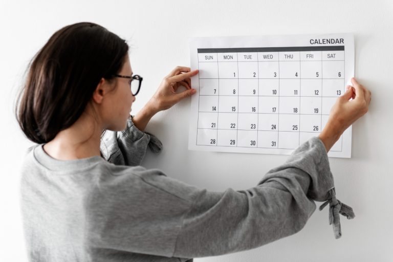 A woman in a gray sweater hangs a well-structured custody calendar on a white wall, adjusting it with both hands.