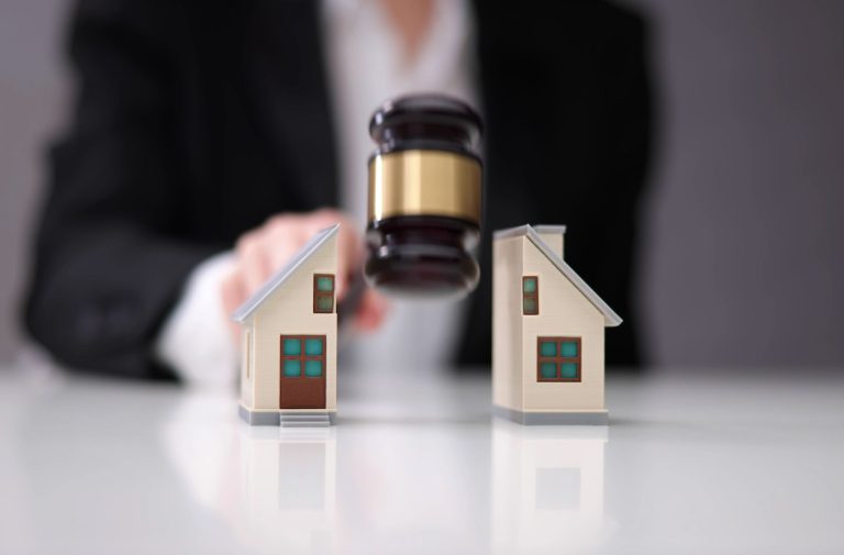 A person in a suit uses a gavel between two model houses, suggesting Houston financial mediators making a legal decision or property division.