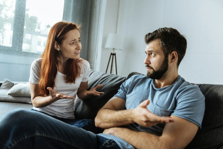 A woman and a man sit on a couch indoors in Houston, facing each other and gesturing with their hands as if having a serious conversation or discussing divorce mediation.