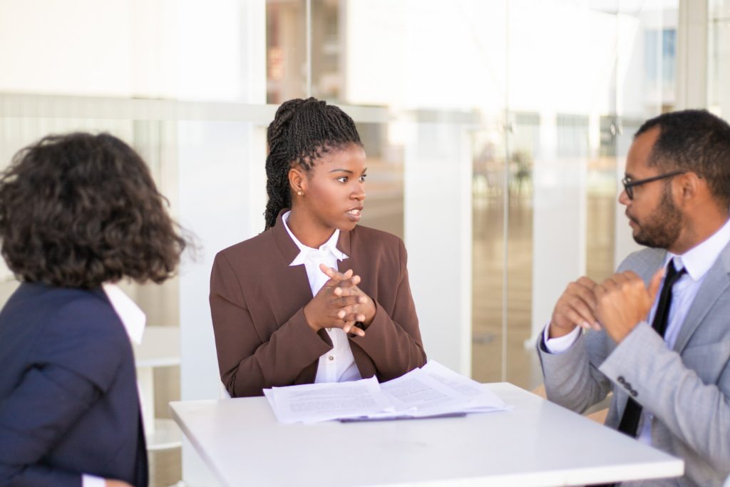 Three Black female divorce attorneys in business attire sit at a table with documents, engaged in a serious discussion in a bright office setting.