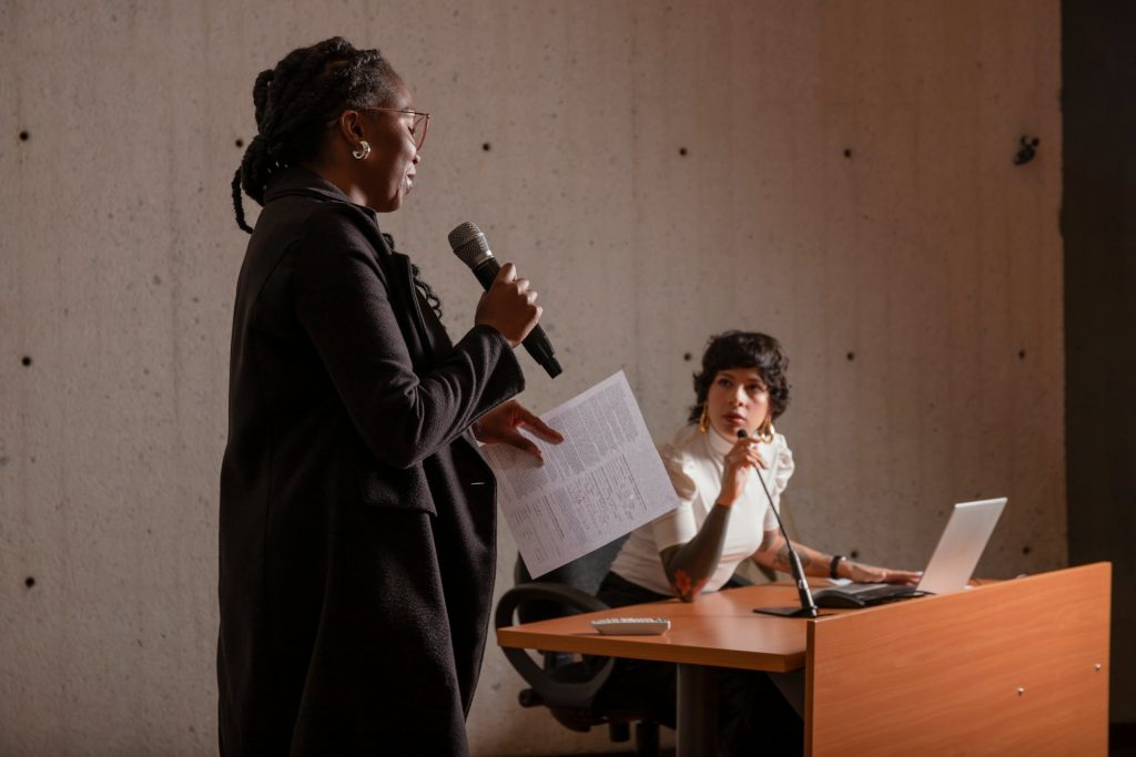 A female lawyer in Houston stands holding a microphone and papers while speaking to another person seated at a desk with a laptop, against a plain wall.