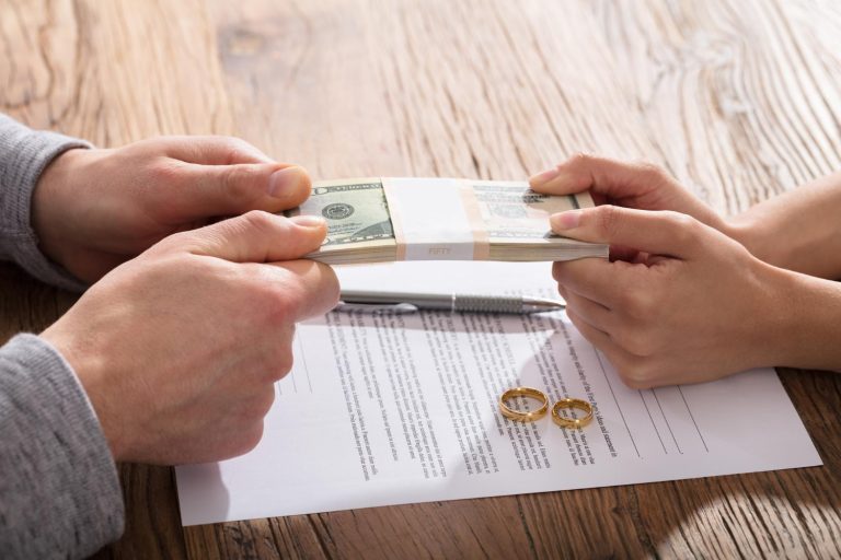 Two people tug on a stack of cash over a document with two wedding rings and a pen, highlighting financial mediation or settlement during divorce proceedings.