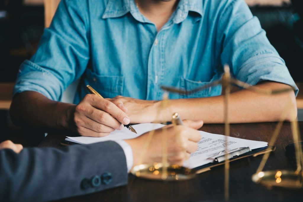 Two people sit at a table reviewing and signing documents, with a pair of golden justice scales in the foreground—a scene reflecting the trusted guidance of Board Certified Family Lawyers.