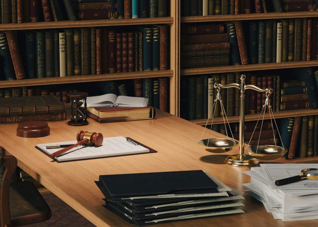 A wooden desk in a law library with a gavel, scales of justice, hourglass, documents, and stacked books—ideal for a Houston family lawyer. Shelves filled with books line the background.