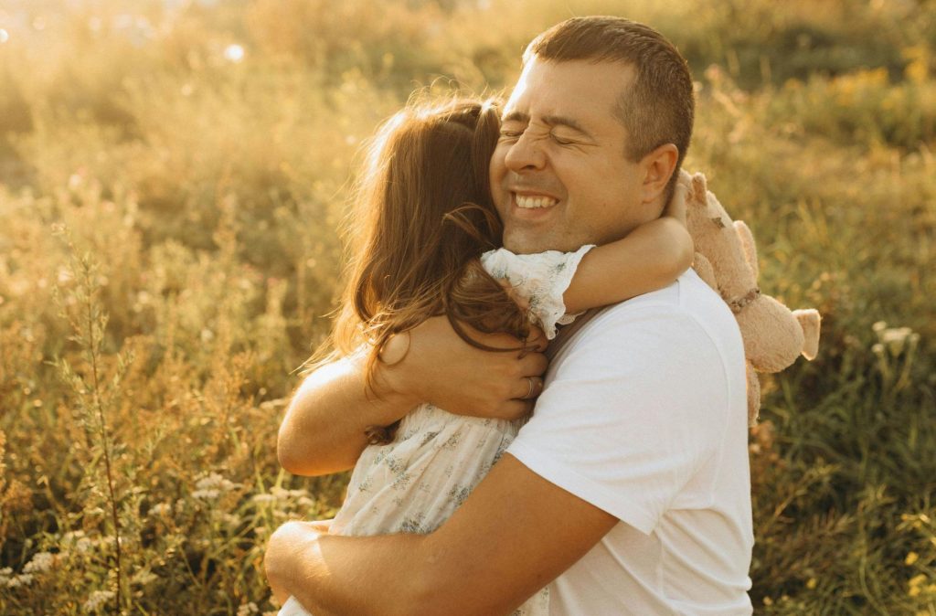 A Board Certified Family Lawyer in a white shirt hugs a young girl holding a stuffed toy outdoors in a sunlit field, both smiling.