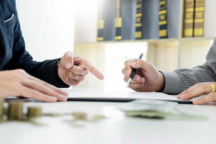 Two people sit at a desk discussing documents, with one pointing and the other signing papers. Coins and bills are visible in the foreground, suggesting a financial mediation process, often crucial in divorce cases.