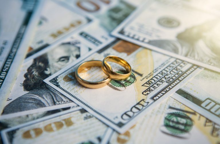 Two gold wedding rings rest atop scattered one hundred dollar bills, symbolizing the financial and emotional complexities tied to visitation schedules and parenting time.