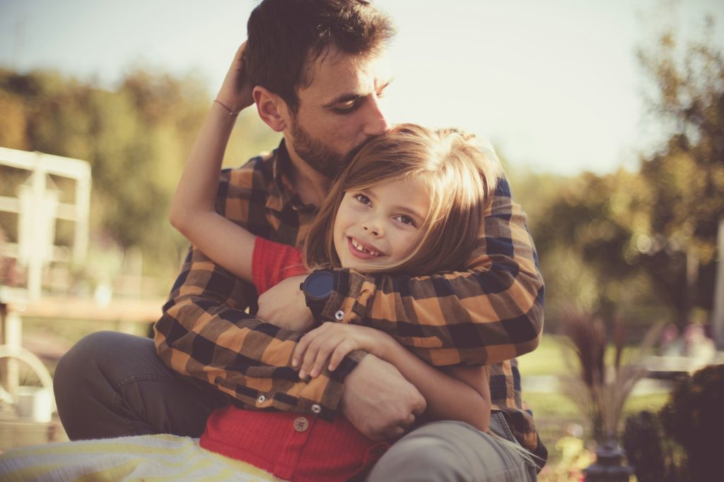 A man in a plaid shirt hugs and kisses a smiling young girl outdoors on a sunny day, capturing a joyful moment that highlights the importance of family during the Texas Child Support process.