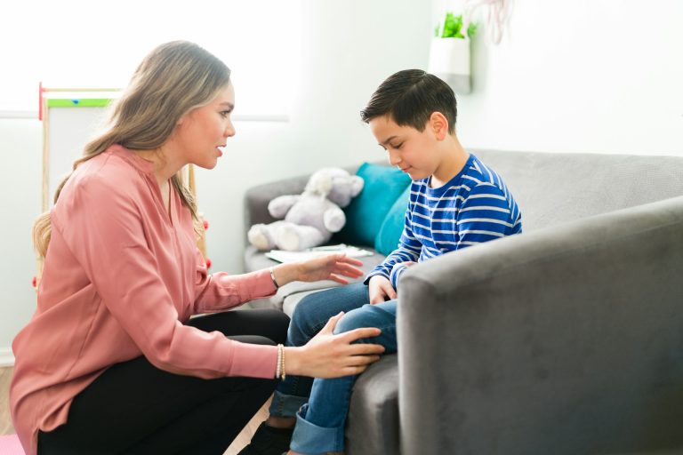 A woman kneels by a sofa, gently talking to a boy in a striped shirt who is sitting and looking down, appearing thoughtful or upset—perhaps discussing concerns about child support hearings or seeking legal advice together.