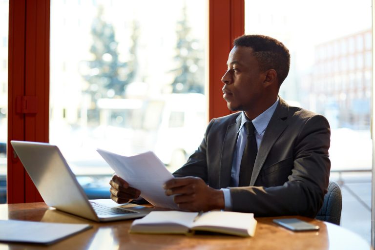 A man in a suit sits at a desk with a laptop, notebook, and phone, holding papers related to divorce as he looks out the window during daytime, possibly considering consultation with financial mediators.
