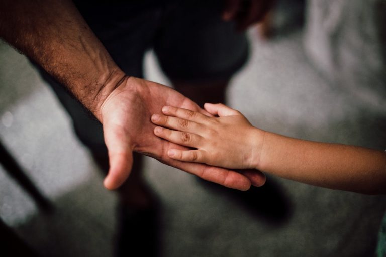 An adult hand and a child's hand touch palm to palm, with the child's hand resting on top of the adult's—a powerful symbol of fathers' rights and the bond often at the heart of child custody disputes.