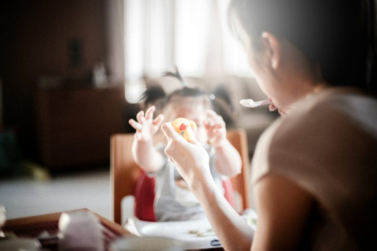 An adult feeds a young child sitting in a high chair, perhaps during their Texas child visitation. The child reaches out with both hands while the adult holds a spoon and a small toy. Natural light fills the room.