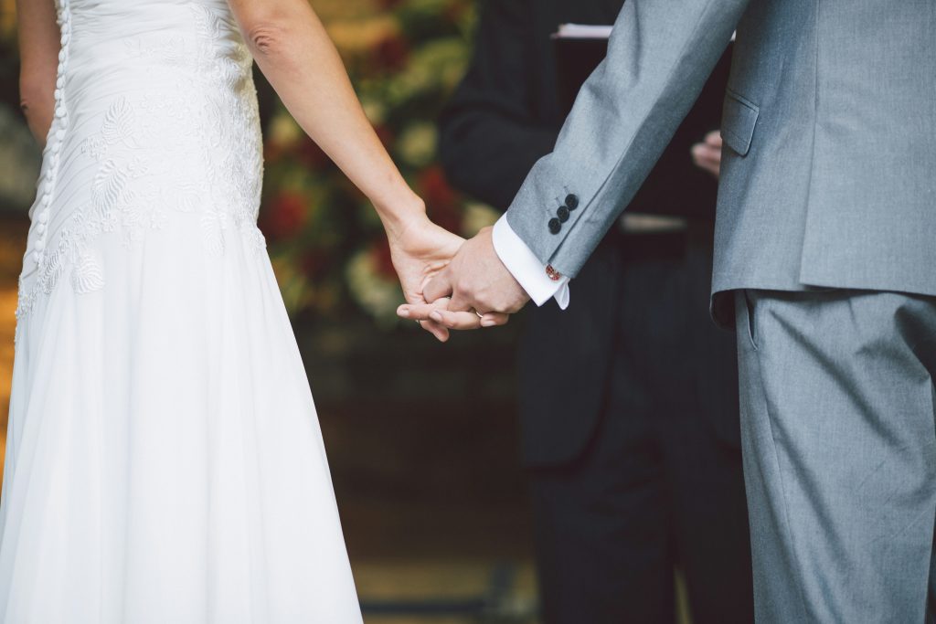 A bride in a white dress and a groom in a gray suit hold hands during their wedding ceremony, ready to start their new life together—backed by the security of a premarital agreement from Angela Faye Brown & Associates.