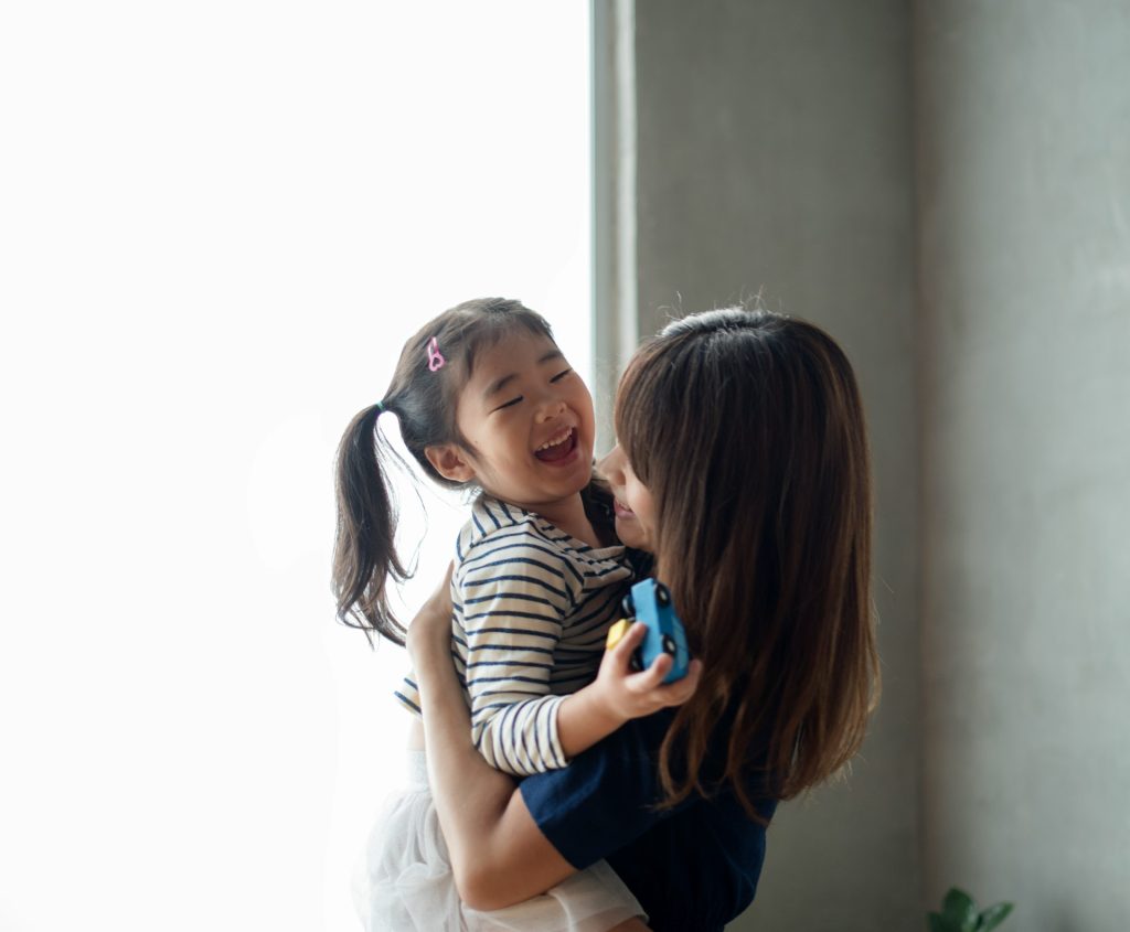 Woman holding and smiling at a young girl with pigtails and a striped shirt; both appear happy in a brightly lit room, enjoying quality time together under the Standard Texas Visitation Schedule.