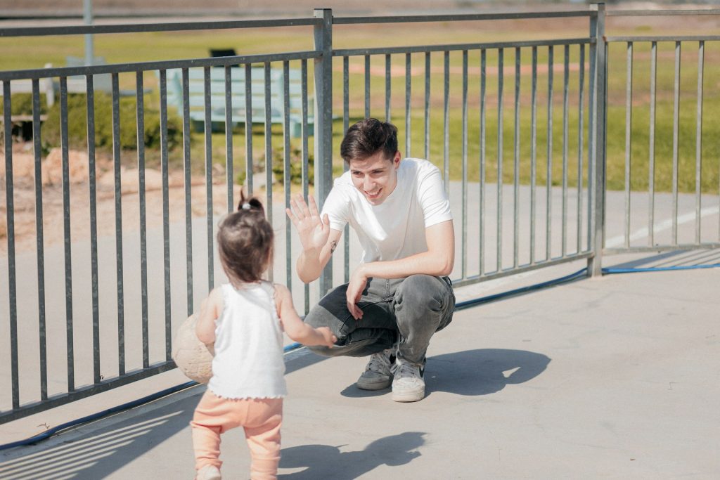 A man crouches and smiles while giving a high-five to a toddler outdoors near a metal fence on a sunny day, capturing the joyful moments that Texas family law aims to protect through visitation rights in Texas.
