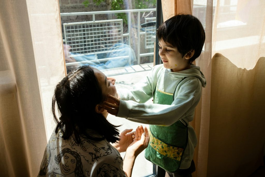 A child in a green hoodie and apron stands by a sunlit window, gently touching an adult's face as they both smile, capturing the warmth and joy at the heart of visitation rights.