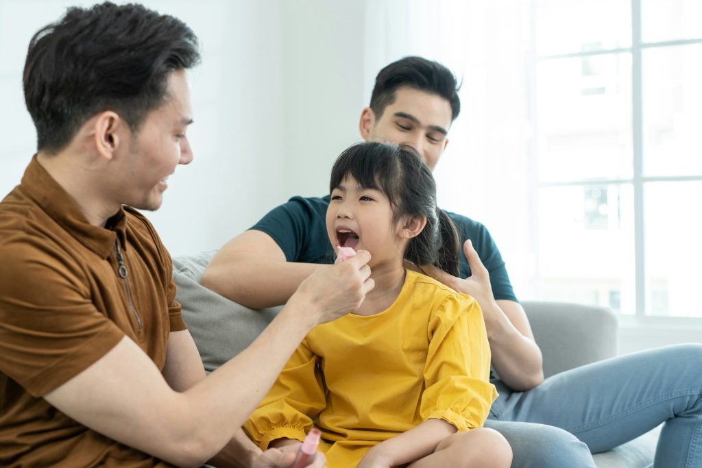A man uses a tongue depressor to examine a young girl’s mouth while another man supports her on a couch in a bright living room, demonstrating care during visitation issues in Texas.