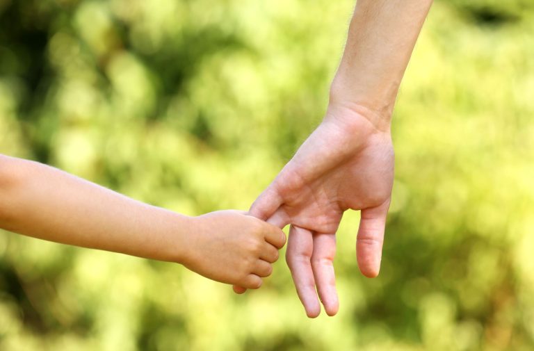 Close-up of a child’s hand gently holding an adult’s finger, with a blurred green outdoor background—a tender moment that echoes the guidance and support families find with a Board Certified Family Lawyer.