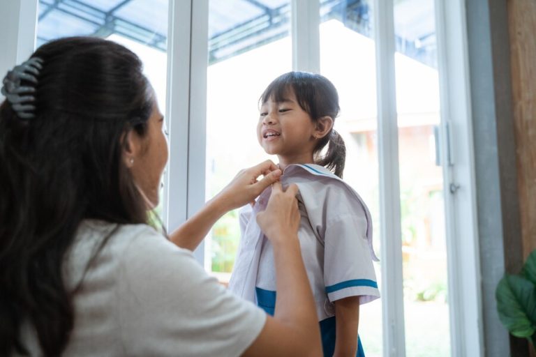 An adult fastens the collar of a young girl's school uniform as she smiles, standing indoors near large glass doors—capturing a tender moment during a child custody visitation in Houston.