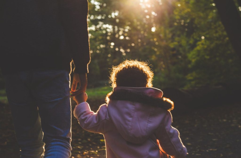 An adult and a child, possibly during parent visitation, walk hand in hand along a sunlit wooded path, with the child in a light pink coat.