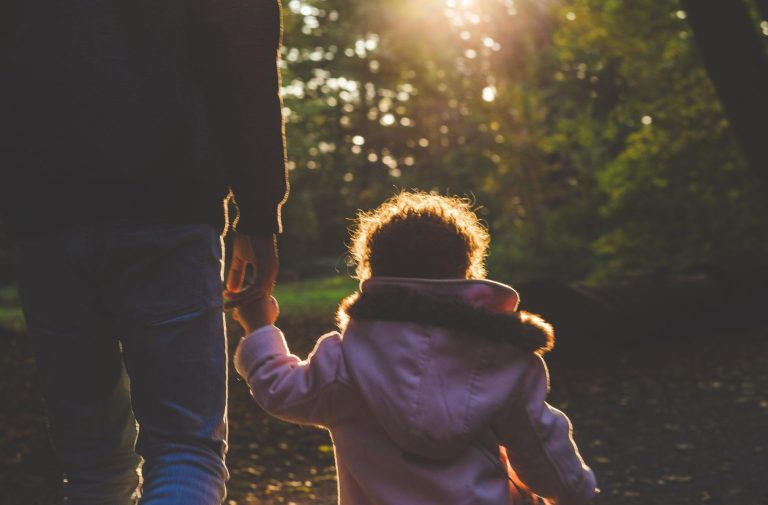 An adult and a child, possibly during parent visitation, walk hand in hand along a sunlit wooded path, with the child in a light pink coat.