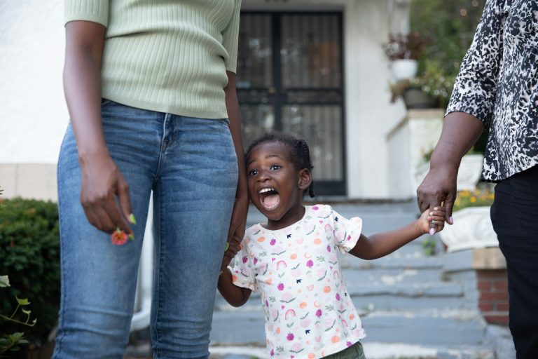 A young child smiles widely while holding hands with two adults, standing outside near steps and a building entrance—capturing a joyful moment during Texas Child Visitation.
