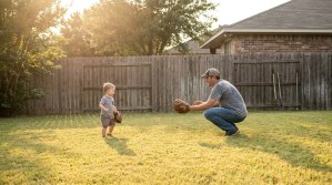 adult man crouching with baseball glove facing toddler wearing mitt in backyard